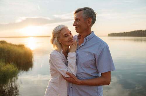A senior couple lovingly embraces by a serene lake during a beautiful sunset.