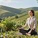 A serene scene capturing a holistic health concept, featuring a balanced individual in professional attire practicing mindfulness on a lush green hillside. In the foreground, show a person of European descent sitting cross-legged, eyes closed, radiating calmness. Surround them with vibrant plants that symbolize healing, like neem leaves and herbs, creating a natural ambiance. In the middle ground, depict gentle soft lighting filtering through trees, enhancing the sense of peace and connection to nature. The background should feature rolling hills under a clear blue sky, conveying tranquility. Ensure the image evokes a sense of harmony between body, mind, and spirit, reflecting a holistic approach to wellness. Use a warm color palette and soft focus for a soothing atmosphere.